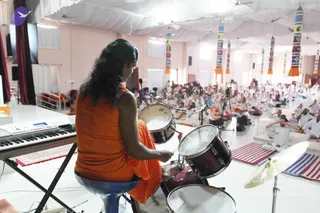 A photo from a large white-draped hall with pink walls and colourful striped pillars, showing a musician seated on a blue stool at a drum kit and keyboard. The drummer, seen from behind, wears an orange sleeveless top and has long dark wavy hair. Beyond, a seated audience of many people in white clothing fills the floor on striped mats, with a camera on a tripod at the right. The scene suggests a satsang or discourse with live music.