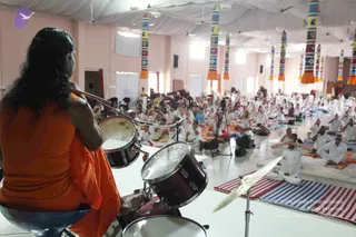 A photograph taken from behind a drummer seated on a stool in the foreground, wearing an orange saffron robe with long dark hair falling down her back, playing a drum kit with mallets. Dozens of devotees in white clothing sit cross-legged on striped mats across a bright pink-walled hall with white draped ceiling and colourful hanging banners. Many attendees have hands folded or are meditating with eyes closed, and a microphone stand and camera tripod stand near the drums. The drummer is the main subject, with the seated white-clad crowd filling the room behind.