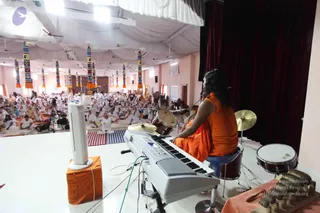 A large hall with white draped ceiling and colourful striped tassel-hung pillars. A seated musician in orange robes with long dark hair sits at a keyboard and drum kit on a raised white stage, facing a crowd of seated devotees in white clothes on white and striped mats. A white floor fan and a small brass lamp stand at the stage edge, with a red curtain on the right. The musician is seen from behind, so the figure is not identifiable from appearance; the scene shows a satsang with musicians and an audience of many white-clad participants.