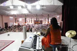A photograph taken from a raised stage looking out over a large hall filled with seated devotees dressed in white. In the foreground, a person with long dark hair wearing an orange sleeveless top and an orange shawl sits on a stool facing the audience, with a keyboard and a drum kit with cymbals beside them. The hall has a white draped ceiling with hanging colourful striped pillars and a red patterned rug at the left edge of the stage. The seated audience fills the floor in rows, many in white clothing, with cameras on tripods visible near the front.