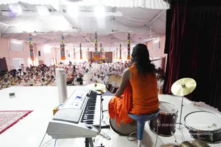 A photograph taken from the stage of a large hall with white draped ceiling and colourful hanging banners. A person seated from behind on a blue stool wears a saffron-orange top and orange wrap, with long dark hair swept down the back, facing a crowd of seated devotees dressed in white. Before them stand a white electronic keyboard, a drum kit with cymbals, a bass drum and a red patterned rug. Many devotees in white clothes sit on the floor in rows, some with a camera on a tripod in the audience area. The seated figure's appearance is consistent with the a person cues of long hair and saffron clothing, though identity remains uncertain from appearance alone.