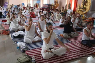 A photograph of a large hall where many people sit cross-legged on striped mats, performing a breathing practice with fingers pressed to their nostrils. Most wear white shirts and white dhoti-style clothing; a few wear dark blue or navy tops. A man in the foreground wears a white shirt, white pants, glasses and a bracelet. Behind them, a golden ornate figure and a gold statue stand near a red curtain, and water bottles sit on the floor. The people are counted as physical attendees in the room.