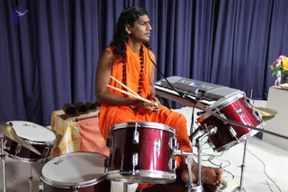 A man in orange saffron robes and a long rudraksha mala sits behind a red drum kit, holding two drumsticks over the drums. Long dark hair falls past his shoulders. A keyboard sits behind him on a white table with a small wooden percussion instrument set and a yellow and pink flower arrangement. A navy blue curtain forms the backdrop, and a microphone stand is nearby. Swamiji (probable): long swept-back hair, orange saffron clothing and rudraksha beads match the page's Swamiji-at-event note, so the person is an appearance-based candidate for Swamiji seated at the drums.