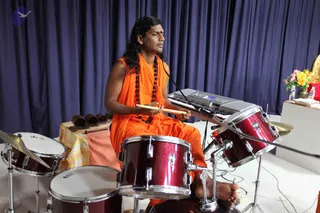 A man in an orange saffron robe and long dark swept-back hair sits barefoot at a drum kit, playing a red drum with a wooden stick while a keyboard sits on a stand behind him. He wears a long rudraksha mala and a red bindi. Blue curtains hang behind the stage, and to the right is a small table with a vase of yellow and pink flowers. Nearby stands a set of bamboo-like wind chimes on a table with a yellow and white checked cloth. Probable Swamiji is visible in the saffron robe, rudraksha mala and long swept-back hair.