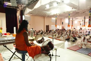 A photograph of a large hall with white draped ceiling fabric and tall cylindrical banners with colourful tassels hanging from the ceiling. In the foreground a figure with long dark hair wearing a flowing saffron-orange robe sits on a stool playing a maroon drum kit, seen from behind. A keyboard stands to the left beside flowers and a small stand. Beyond, a seated audience of many people, mostly dressed in white, sits on mats and cushions facing the stage, with some on striped and blue mats at the right and a man in white seated near the front right.