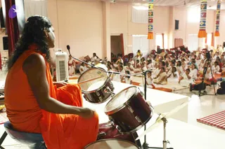 A photograph of a long-haired person in an orange saffron robe seated on a stool at a drum kit, playing a snare and tom with drumsticks on a raised white stage. A large audience of people in white clothing sits cross-legged on mats in a pastel-pink hall, facing the stage. Two colourful woven pennants hang from the upper walls, and a camera on a tripod records the event. A white fan stands near the drums. The person's appearance is consistent with a person candidate, with long swept-back hair and orange saffron clothing, but this is uncertain.