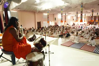 A photograph of a hall-wide satsang seen from the stage. In the foreground a person with long dark hair sits on a stool at a drum kit, dressed in a flowing saffron-orange robe, holding a drumstick and a microphone on a stand. Facing the stage, a large audience in white clothing sits cross-legged on striped and plain mats across the floor. Tall hanging pillars with colourful patterned bands and orange tassels line the pale pink hall beneath white draped ceiling fabric. a person is not clearly identifiable from appearance, so the drummer is described by role only.