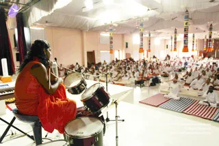 A photograph taken from behind a stage shows a person with long dark hair seated on a stool in a saffron-orange garment, playing a set of drums with sticks in the foreground. Nearby stand a keyboard and microphone stands. Beyond the stage, a large hall is filled with hundreds of seated devotees dressed in white, sitting on striped and blue-patterned mats. The ceiling is draped in white fabric and hung with tall decorative pillars in red, blue and white patterns with orange tassels.