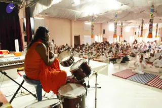 A photograph of a large hall with white draped ceiling and colourful striped hanging banners. In the foreground a person with long dark hair wearing an orange robe sits on a stool at a drum kit, holding a microphone and looking toward the audience. A keyboard stands to the left. Hundreds of seated attendees in white clothes fill the floor on striped mats, with a camera on a tripod at right.