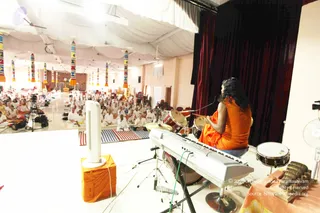A wide photograph of a discourse hall seen from the stage. In the foreground a person in an orange saffron robe with long dark hair sits on a low seat, back to the camera, facing a large seated congregation. Behind the seated audience, many people in white clothes sit on striped mats and floor cushions across the hall. White ceiling drapes hang overhead, with colourful striped pillar banners lining the room. The stage holds a keyboard, drum kit, a tall white fan, an orange-covered stool and a tabla-style drum. A camera on a tripod films the event.