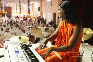 A photograph of a performer seated at a drum kit on a stage, playing drumsticks on a snare drum with a keyboard in the foreground. He wears an orange saffron robe draped over the shoulders, a dark brown rudraksha mala around the neck, and has long, swept-back dark hair; he looks toward the audience. Behind him are cymbals and a bass drum. In the hall, a large seated crowd of people in white clothing sits on the floor facing the stage, beneath hanging colourful banners and pink walls. Swamiji (probable) based on the long hair, saffron robe and rudraksha mala, playing music on stage before the seated devotees.