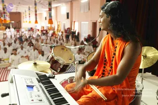 A person with long dark wavy hair wears an orange-saffron sleeveless garment with a matching orange wrap draped over the shoulders and a dark rudraksha mala around the neck. Seated at a drum kit with drumsticks in hand, the person plays a bongo-style drum set beside a keyboard in the foreground. Behind, a large hall holds a seated audience of people dressed in white, with cymbals, a drum kit and colourful decorative pillars and banners overhead. The person's appearance with long hair, saffron clothing and rudraksha beads is consistent with Swamiji (probable), seated as a musician during a satsang session.