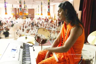 A photograph of a long-haired figure in a sleeveless orange robe with a matching orange shawl and a dark rudraksha mala, seated at a drum kit with drumsticks in hand beside a keyboard. Behind them, a large hall is filled with rows of seated devotees dressed in white. Colourful tasselled hangings decorate the pillars and a golden figure stands in the background. Swamiji (probable) is the person playing the drums, based on the long hair, orange clothing and rudraksha mala.