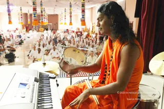 A performer in a sleeveless orange-saffron robe with a long, swept-back dark hairstyle and a rudraksha mala sits at a drum kit, playing a small tabla-style drum held in one hand while a keyboard sits in the foreground. Behind him a large hall is filled with seated devotees in white clothing, and colourful striped tassel hangings and draped white fabric hang from the ceiling. The appearance and long saffron robe with rudraksha beads match the cues for a person, shown here as a musician leading the music, but the image is uncertain. Several white-clad devotees are seated on the floor facing the stage.