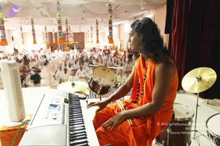 A person in a saffron-orange sleeveless garment and matching shawl, with a dark rudraksha-style bead mala around the neck, sits at a keyboard on the left and plays drums on the right. Long dark hair falls past the shoulders. Behind the stage, a large hall with white ceiling drapes and colourful tassel decorations holds a seated audience of many people dressed mostly in white. A red curtain hangs at the right edge, and a standing lamp and camera tripod are visible in the foreground. The player's appearance is consistent with the a person cues described in the page, but the person's identity cannot be confirmed from appearance alone.