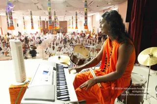 A person in an orange-saffron robe with dark curly shoulder-length hair and a rudraksha-style mala sits at a keyboard on a white stage, with drumsticks in hand and a drum kit beside them. In front of the stage, a large seated audience in white clothes fills a hall under a white draped ceiling hung with colourful striped pillar banners. A deity murti and a small golden figure are visible at the far end near the stage.
