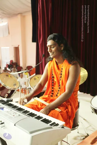 A man in orange saffron robes and a long rudraksha mala sits at a white electronic keyboard, playing it with his right hand while holding a pair of drumsticks in his left, on a stage with a deep maroon curtain behind him. Two drums and a cymbal are visible behind him, and a microphone arm extends toward him. Several seated people in white clothes are visible in the blurred background at the left. The man has long, swept-back dark hair and a red bindi on his forehead; the page's a person flag is not enough to establish his identity from appearance, so he is described by role as a performer.