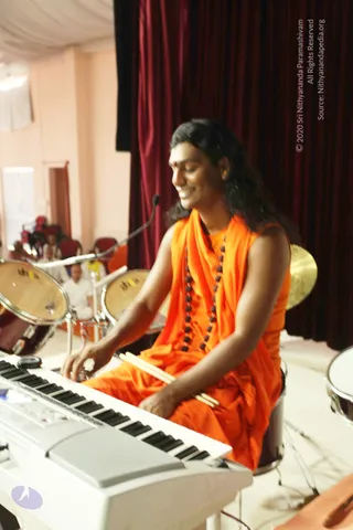 A smiling man in an orange saffron robe with a rudraksha mala sits playing a white electronic keyboard on a stage in front of dark red curtains. He wears a red bindi and has long dark hair, and his hands are on the keys with drumsticks in his right hand. Behind him are drum kits and a cymbal, and a few seated people in white clothes are visible in the background hall.
