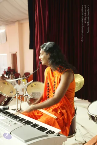 A performer in a flowing orange saffron garment with a rudraksha mala draped over the chest sits at a white electronic keyboard, playing drumsticks on a drum kit on a stage. Long dark hair sweeps back, and a microphone stands in front of him. Behind is a deep red velvet curtain, with a gold cymbal visible. In the background, a few seated audience members in white clothes watch from red chairs in a pale pink hall.