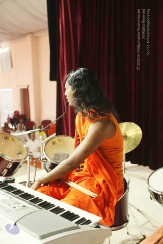 A figure in an orange saffron robe with long curly black hair and a rudraksha mala sits on a drum stool at a white keyboard, holding drumsticks over the keys while speaking into a microphone on a stand. Behind them are a red velvet curtain, a tom drum and a cymbal on a stage. In the background, several seated people in white clothes and red chairs face the stage, and a person candidate is the person in orange performing as a musician.
