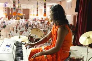 A photograph taken from behind a performer seated on a stool on a stage, playing a drum kit and keyboard. The performer wears an orange-saffron sleeveless garment draped over the body and a dark rudraksha-style bead necklace, with long dark wavy hair falling past the shoulders; the performer is shown in profile facing the audience. A red-and-white striped drum rests before them, a bass drum labelled with a brand name sits near the keyboard, and two cymbals are visible. Beyond the stage a large hall holds a seated audience of people dressed largely in white, arranged in rows toward the back. Colourful vertical striped pillars with orange tassels hang from the ceiling, and a red curtain is at the right edge.