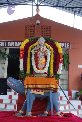 A photograph of a Ganesha murti standing on a grey mouse vehicle (vahana) in front of a temple building with a red-orange wall. The Ganesha figure is decorated with a silver crown, jewelled ornaments, a red shoulder cloth and an orange dhoti, and sits within a rainbow-shaped arch of marigold, rose and jasmine garlands with green leaves. The mouse has a red-and-white painted mouth and an orange cloth draped over its back. Striped red-and-white steps lead up to the entrance, and a red cloth covers the ground. A signboard on the wall reads Sri Ananda ... Temple. No people are visible.