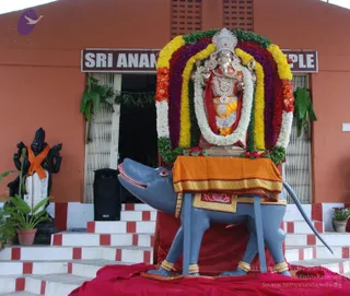 Outdoor photograph of a temple entrance with a salmon-orange wall and a dark brown signboard reading "SRI ANAN... TEMPLE", partly cut off. A large Ganesha murti sits on a grey mouse vahana (vehicle) on a red-cloth-draped platform at the top of white and red steps. The murti is decorated with a dense arch of marigold and rose garlands in red, orange, yellow, purple and white, a silver crown, and an orange-gold cloth draped over the mouse. A black statue of a standing deity in white dhoti with an orange shawl stands to the left, with potted plants nearby. No people are visible.