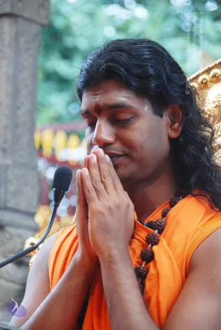A man with long, swept-back dark hair sits with eyes closed and palms pressed together in namaste at chest height. He wears a sleeveless saffron-orange garment draped over the shoulders and a long rudraksha mala of brown beads. A red kumkum mark and white vibhuti line are on his forehead. A small microphone on a flexible stand is at left, and a golden ornate chair or throne edge appears at right against green foliage and a stone pillar. This appearance-based label applies: Swamiji (probable), with long hair, saffron clothing and rudraksha cues, seated in a discourse setting.