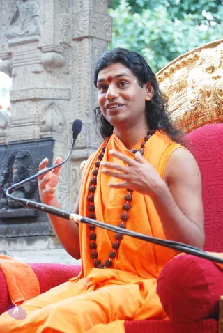 A seated man in an orange sleeveless top and orange wrap speaks into a small clip microphone on a boom stand, his hands raised mid-gesture. Long dark hair falls past his shoulders, and a red bindi marks his forehead. He wears a long rudraksha mala. He sits on a red upholstered chair with an ornate gold carved backrest. Behind him is a weathered stone pillar carved with figures, and a dark stone deity relief stands at left beside green foliage. Appearance-based label: Swamiji (probable), delivering a discourse, identified by the long hair, orange garments and rudraksha mala cues.