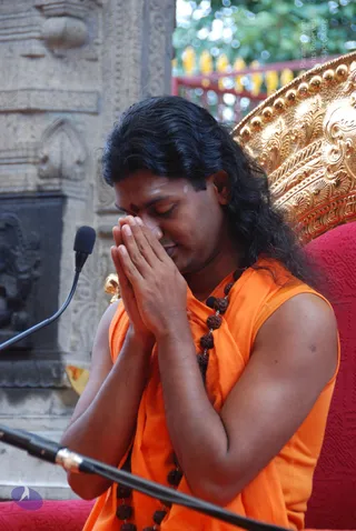 A man with long, swept-back dark hair and a small red tilak on his forehead sits with palms pressed together in anjali mudra, eyes lowered, in front of a microphone. He wears a sleeveless saffron-orange garment and a long rudraksha mala with dark beads. Behind him is an ornate gold throne-back with a red cushion, beside a carved stone pillar with relief sculpture.