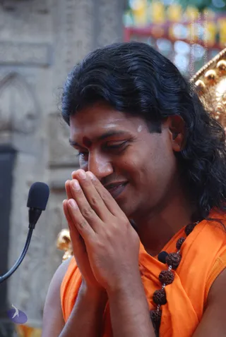 A man with long dark wavy hair and a red bindi on his forehead, wearing an orange sleeveless garment and a strand of rudraksha beads, sits with palms pressed together in anjali mudra, eyes lowered and smiling. A small microphone on a flexible stand is at left, a golden carved throne edge is at right, and a stone carved pillar stands behind. Probable Swamiji: long swept-back hair, orange clothing and rudraksha mala are visible, so the candidate is shown in person in a discourse setting.