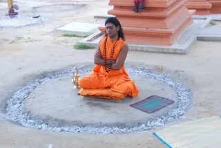 A photograph of a man seated cross-legged on a raised concrete platform inside a circle of white ash outdoors. He wears an orange saffron robe, a long rudraksha mala around his neck, and a red tilak on his forehead; his long dark hair falls past his shoulders and his arms are folded across his chest with eyes closed in meditation. A brass kalasha-style vessel sits beside him on the platform, a small patterned mat lies on the ground nearby, and an orange tiered shrine base with a garland of flowers stands behind him. Dry earth and a sparse patch of grass surround the scene.