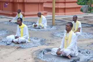 Five men sit cross-legged in meditation on a sandy, ash-dusted courtyard in front of orange-red temple walls. All wear white dhotis and white shirts with yellow shawls draped over their shoulders. Their eyes are closed or lowered and their hands rest in their laps; one man in the foreground holds his hands folded. Each sits on a small mat or cloth placed on a patch of grey ash, which appears to mark where fire pits once stood. A yellow-painted wooden pole stands among them, and a few plants grow at the edge of the frame.