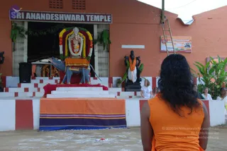 A photograph of Sri Anandeshwara Temple, with a long-haired figure seen from behind in an orange sleeveless top standing before a shrine on a sandy courtyard. The shrine holds a deity murti in a garland of yellow, orange and purple flowers, mounted on a grey bull mount (nandi) with an orange cloth draped over it, with a dark Shiva-type sculpture beside it. Two women in white stand at the side, and a few more people are partly visible at the right edge. The wall has a painted sign reading Sri Anandeshwara Temple, and a bamboo ladder leans against the orange wall. Swamiji (probable) is the long-haired figure in orange, identified by long swept-back hair and saffron clothing.