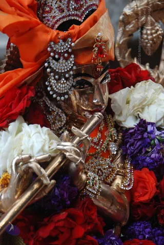 Close photograph of a golden metal deity murti playing a long flute held to its lips, decorated with an orange silk turban crowned with jewelled ornaments, pearl and crystal earrings, and layered jewelled necklaces. The murti is surrounded by red roses, white roses and purple flowers, with a golden ornamental pillar in the upper right.