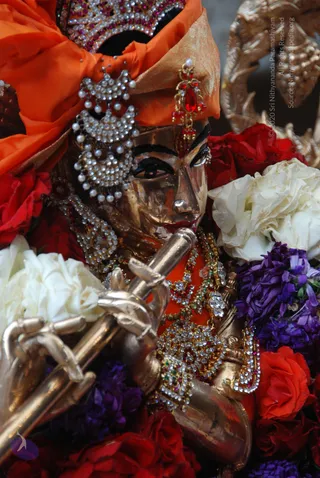 Close-up of a golden metal murti of a deity playing a long flute, its face shown in profile with dark kohl-lined eyes and red lips. The deity wears an orange turban topped with a jewelled crown, dangling pearl-and-stone earrings, and layered necklaces set with red and green stones. Red roses, white roses and purple flowers surround the figure, and an ornate golden backdrop sits at the upper right. No people are visible; the image shows only the statue and flowers.