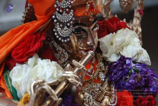 Close-up photograph of a gilded murti of Krishna playing a golden flute, its face painted in metallic gold with dark eyes. The figure wears an orange turban-style headdress with a jeweled crown, pearl and rhinestone ornaments, and layered necklaces with red and green stones. Red roses, white roses and purple blossoms are heaped around the image, and an orange cloth drapes over the left side. No people are visible; the subject is a deity statue.