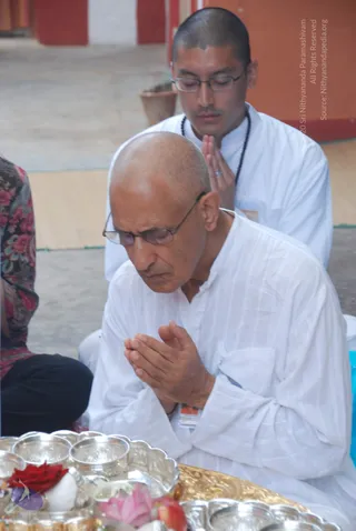 A photograph of two men seated on the floor in white ritual clothing, hands pressed together in anjali mudra with eyes closed. The man in front is bald and wears wire-rimmed glasses and a plain white kurta. Behind him sits a younger man with a shaved head, glasses and a dark rudraksha mala over a white shirt. In the foreground are silver plates holding a red flower, a pink flower and a white offering, with a gold tray beneath them. A patterned sari or fabric edge appears at the left edge of the frame, and a red wall is visible in the background. No one in the scene wears saffron or ornate a person-style clothing, so no identity candidate is claimed.