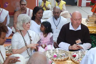 A seated group during a puja in an outdoor courtyard. In the foreground, a woman with short silver-white hair wears a white sari, a rudraksha mala and bangles, and holds her hands together with a small girl in a pink dress and gold necklace beside her. Behind them, a shaved-headed man in white with a tilak and beaded mala sits next to a woman in white. On the right, a bald man in a brown sweater over white clothes, wearing an ID badge, sits with folded hands before a gold tray. Silver and brass plates of flowers, rice and offerings sit on a golden tray, with red and pink flowers in front. Other devotees in white and yellow sit behind, and a gilded altar stand appears at the upper right.