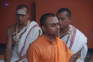 Three men sit in a row before a red-orange wall during a puja. The central man, eyes closed, wears a saffron-orange robe and has white tripundra lines and a red bindi on his forehead. On the left, a bespectacled man in white dhoti and shawl with maroon borders, wearing gold beaded necklaces and white ash markings on his forehead and arms, looks down. On the right, another man in a white dhoti with maroon borders and gold beaded necklace, with a red mark and white lines on his forehead, sits close behind the central figure. No figure in the frame has distinctive a person cues such as long hair, crown, rudraksha mala or ceremonial gold necklace beyond the ordinary gold beads worn by the priests, so identity is uncertain.
