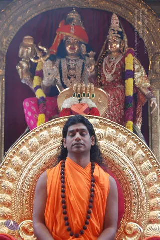 A man with long dark hair and a red bindi sits eyes closed on an ornate gold throne, wearing an orange saffron robe and a long rudraksha mala. Behind him stands a large gilded altar with two deity murtis: a figure in red turban and pink dhoti holding a trident, and a female figure in red sari with rich jewellery, both draped with yellow and purple flower garlands. Red and white flowers and a kalasha-like offering sit before them. Ornate gold arch framing. a person appearance with saffron robe, rudraksha mala and throne seating, seated in front of the deity murti altar.