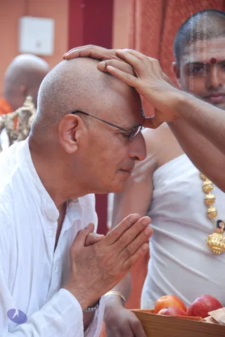 A close photograph of a shaven-headed man in white clothes, wearing thin-framed glasses, kneeling or bowing with palms pressed together in namaste. Another man in white clothes with a red tilak on his forehead and a gold necklace with a small framed portrait pendant places his hand on the bowed man's head. A tray of red apples and other offerings sits in the lower right, with an orange-robed figure partially visible behind. The scene suggests a blessing or honouring ritual.