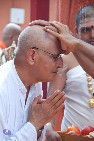 A photograph of a shaved-headed man in white kurta with thin-framed glasses, eyes closed, palms pressed together in namaskar, bowing his head. Another person in white dhoti and gold necklace, with a red tilak on the forehead, places a hand on the bowed man's head, a blessing gesture. A wooden tray of red apples sits at the lower right, and an orange cloth backdrop is behind them. Other people are partly visible at the edges.