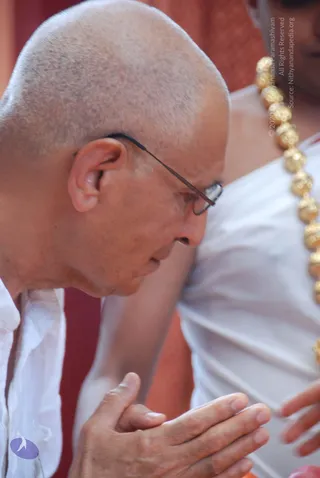 Close-up photograph of a shaved-headed man in thin wire-rimmed glasses and a plain white shirt, head bowed and palms pressed together in anjali mudra. Beside him, a person in white clothing wears a long strand of large gold beads. The background is a warm red-orange fabric.