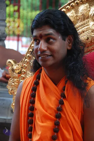 A close-up photograph of a man with long, dark, wavy hair wearing an orange saffron shawl over his shoulders and a long strand of dark brown rudraksha beads across his chest. He has a red bindi and white tilak on his forehead and sits in front of an ornate gold throne with round gold knobs and carved scrollwork. Behind him is a red cushion and part of a green-and-red background. He looks to the side and smiles slightly. Swamiji (probable): long swept-back hair, saffron shawl and rudraksha mala visible; the image is captioned on the page as Sarva Darsan, and the figure is seated on a golden throne.