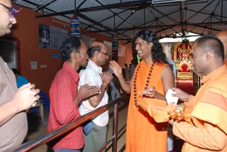 A photograph inside a covered hall with orange-painted walls and a corrugated roof. A long-haired man in a saffron-orange robe and dark rudraksha mala smiles and gestures with raised palm toward a row of visitors waiting along a metal railing. On the left, a man in a red shirt and a man in a white striped shirt press their palms together in greeting; a man in a brown shirt stands at the edge. On the right, a young man in orange robes holds a small gold-coloured vessel and white paper. Behind them, a richly decorated shrine with gold and red ornaments holds a deity murti adorned with garlands, and a wall of framed posters and printed notices is visible on the left.