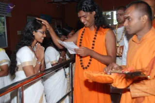 A photograph of a sannyasi in orange robes with long swept-back dark hair and a dark rudraksha mala standing at a railing, placing his hand on the forehead of a smiling young woman in white traditional dress with gold borders who has her palms pressed together in namaste. Behind him an orange-robed monk holds a tray with orange cloth and a lit oil lamp-like object, and another man in white with a gold chain stands nearby. Several women in white saris with gold borders stand in the background. The setting is an indoor hall with a red wall and a window. The robed man with long hair and malas is a person by appearance cues, shown giving blessing in a ritual setting.