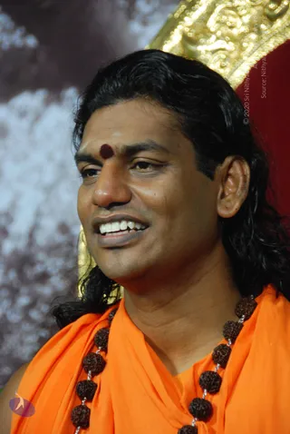 Close photo of a smiling man with long dark hair swept back, wearing an orange saffron shawl and a multi-strand rudraksha mala with large brown beads. A round red-brown bindi and white vertical tilak mark his forehead. A gold ornate throne or canopy edge is visible behind him against a cloudy blue-grey backdrop.