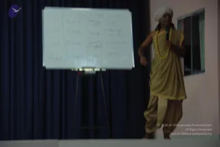A blurred photograph of a speaker on a low stage giving a talk with a handheld microphone. He wears a white turban, a golden-beige unstitched robe draped over one shoulder with matching beige dhoti-style trousers, and a yellow beaded necklace. Behind him is a whiteboard on an easel with handwritten notes in columns, in front of a dark blue curtain. A window and pale wall are visible on the right. The speaker's identity is uncertain from appearance; the setting suggests a discourse session.
