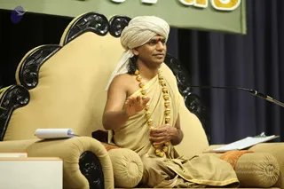 A photograph of a man seated on an ornate, cream-upholstered carved wooden throne with black scrollwork, speaking with his right hand raised in a gesture. He wears a cream turban with a red bindi on the forehead, a beige-gold draped robe with gold trim, and a long necklace of large gold beads. A microphone on a stand is at right, a notepad rests on the armrest, and a dark blue curtain and green banner with partial lettering form the stage backdrop. a person: the cues are the turban, gold bead necklace and ceremonial robe, seated as the speaker on the throne at a discourse.